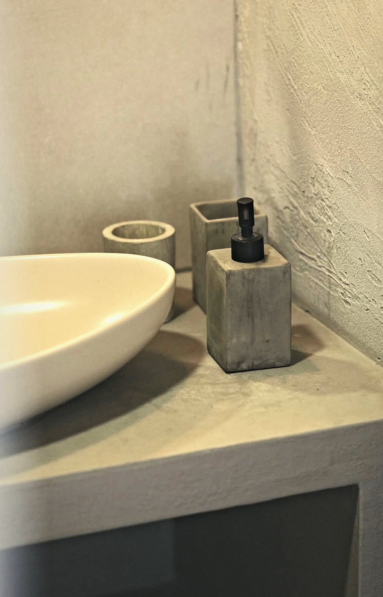 An oval cream basin with concrete soap dispenser and cups on a plastered vanity