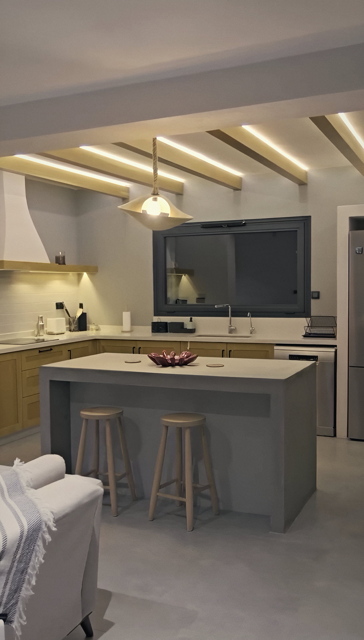 Open kitchen with oak cabinets, a plastered island, two wooden stools and lit ceiling beams