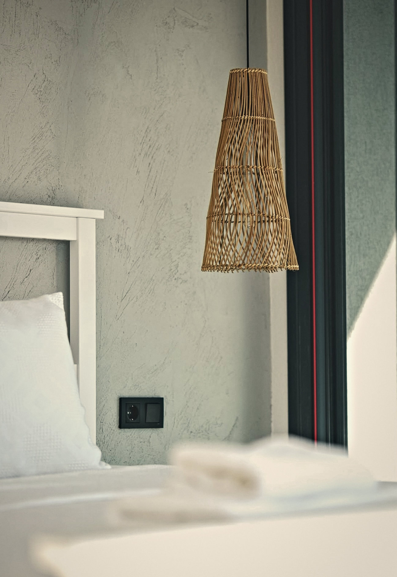 A rattan pendant beside a white bed against a textured plaster wall