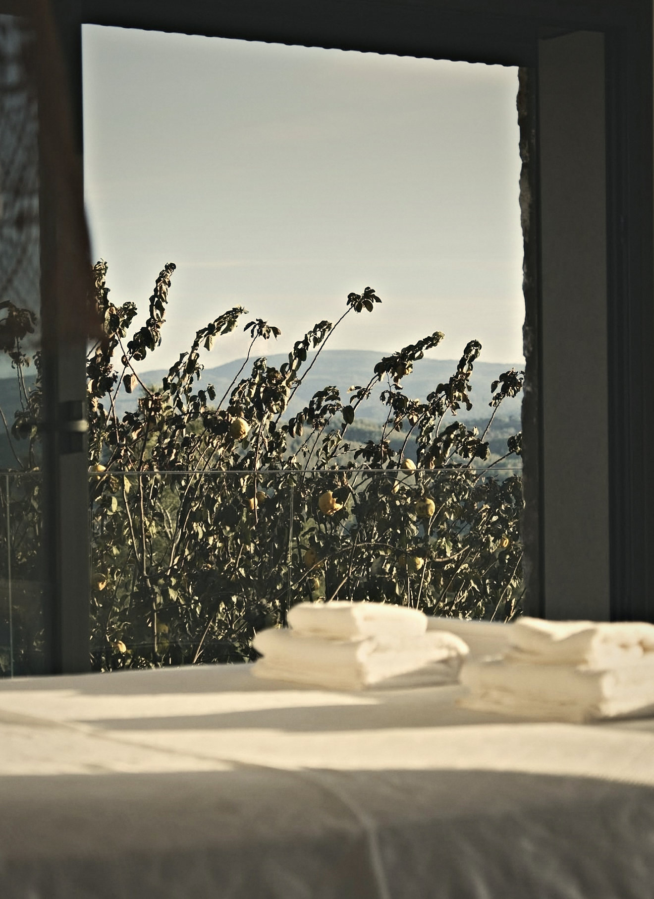 Folded white towels on a bed in morning sun, the garden and hills through the window